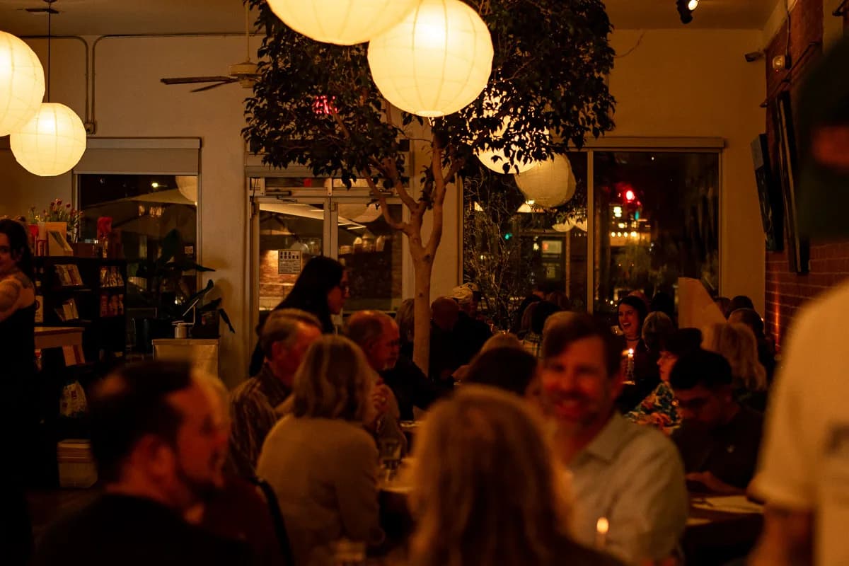 A full room at a Tucson Foodie member dinner: guests talking and laughing along long tables under paper lanterns.