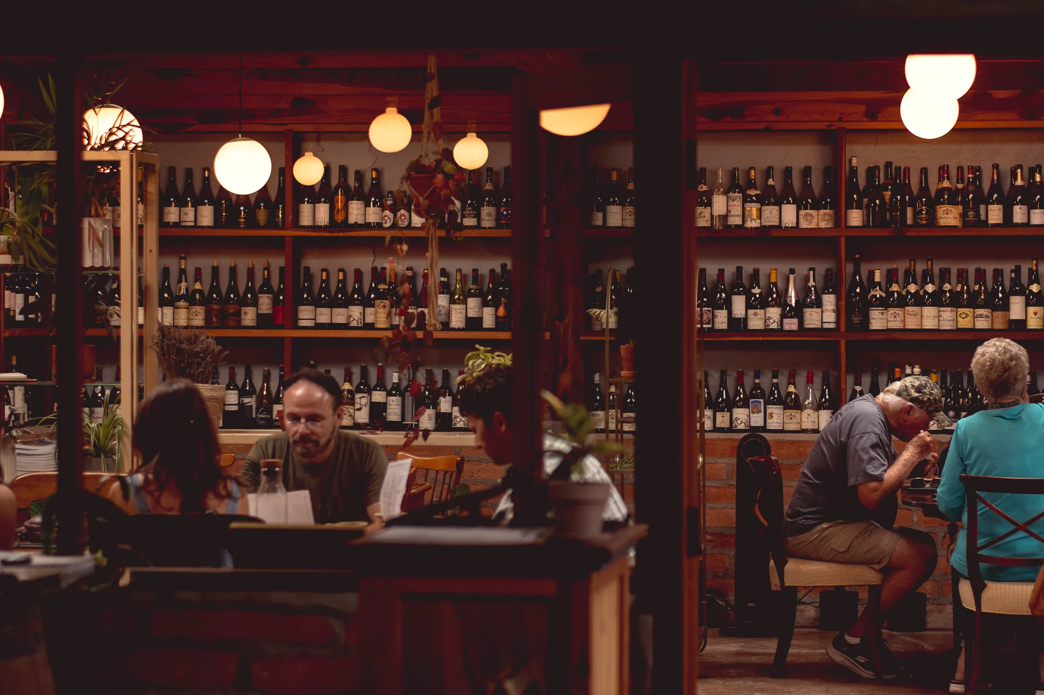 Guests at tables in a warmly lit Tucson dining room, floor-to-ceiling shelves of wine bottles behind them under globe pendant lights.