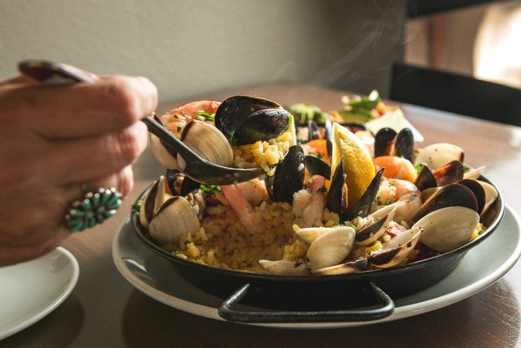 A person with light-medium skin tone, wearing a turquoise ring, uses a dark-colored spoon to lift a portion of a seafood paella from a black, shallow, round pan. The paella consists of yellow rice, mussels, clams, shrimp, and a lemon wedge. Steam rises from the dish. The pan sits on a white plate, which is on a wooden table. A partially visible white plate is in the lower left corner. The background is blurred but shows a portion of a wooden table and a dark-colored chair. The lighting is warm and highlights the food.