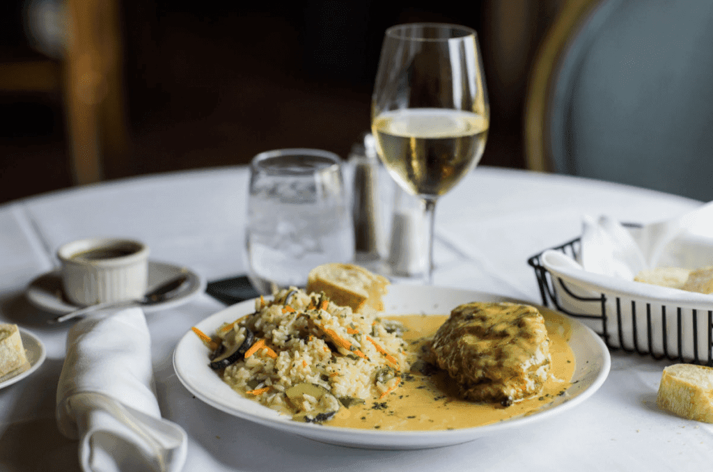 A white tablecloth covers a round table set for a meal. A white plate holds a portion of rice pilaf with mixed vegetables (zucchini, carrots, and possibly green beans) in a creamy sauce. Next to the rice is a piece of breaded and pan-fried chicken breast, also covered in the same creamy sauce. Garnished with fresh herbs. A glass of white wine sits to the right of the plate. To the left, there is a small white ramekin containing a dark green sauce, a folded white napkin, and a piece of bread on a small side plate. A glass of ice water is partially visible behind the plate. More bread is visible in a wire bread basket in the background. The background is blurred but shows a dark interior with hints of wood tones. The lighting is soft and warm, focusing on the food.