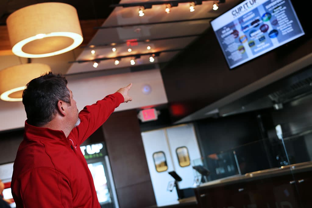 Cup It Up owner Chris Smith motions to one of the 50 inch smart television menu screens. (Credit: Adam Lehrman)