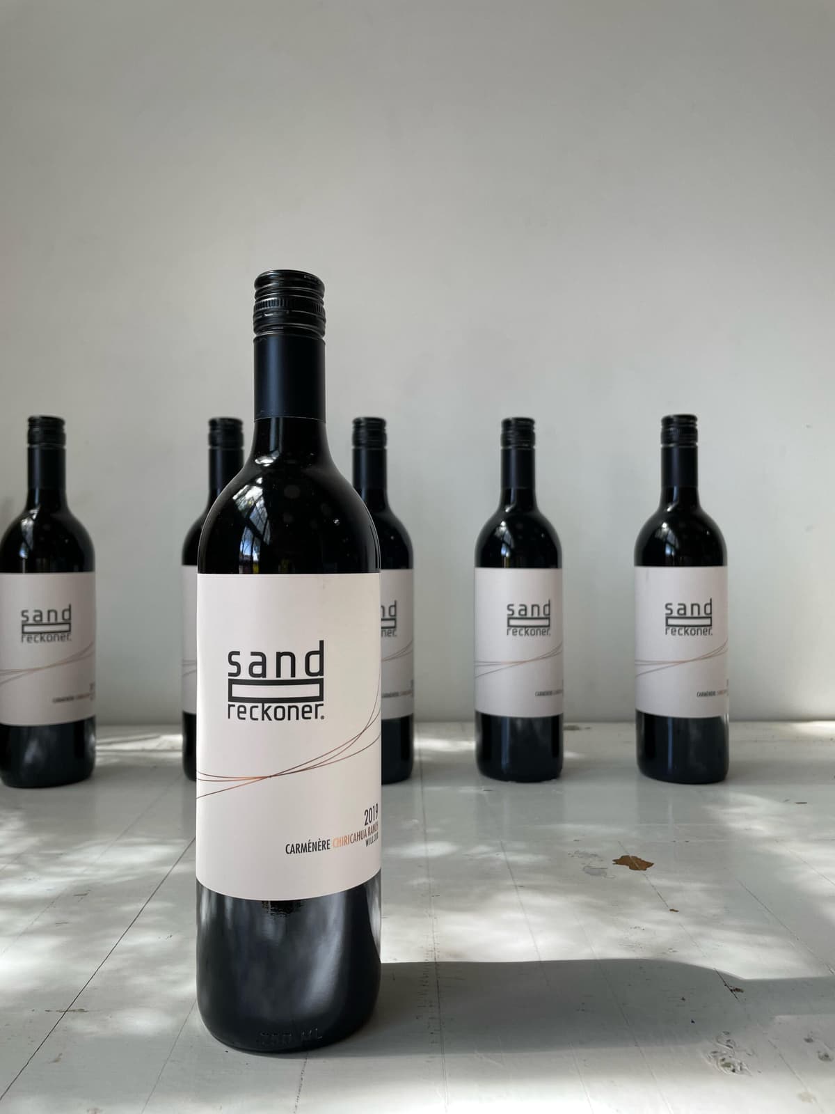 A bottle of Sand-Reckoner 2019 Carmenere in front of a row of matching bottles on a sunlit white table