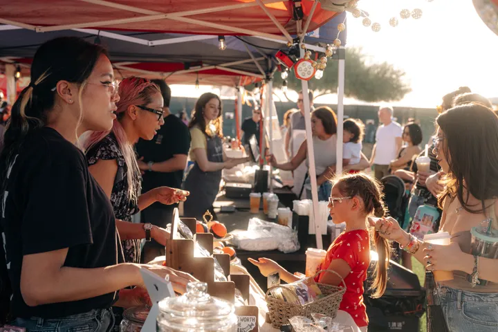 Shoppers at a food stall under a red tent as the sun sets at the May 2026 market.