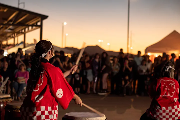 A taiko drummer in a red happi coat mid-strike, a crowd and the sunset sky behind her.